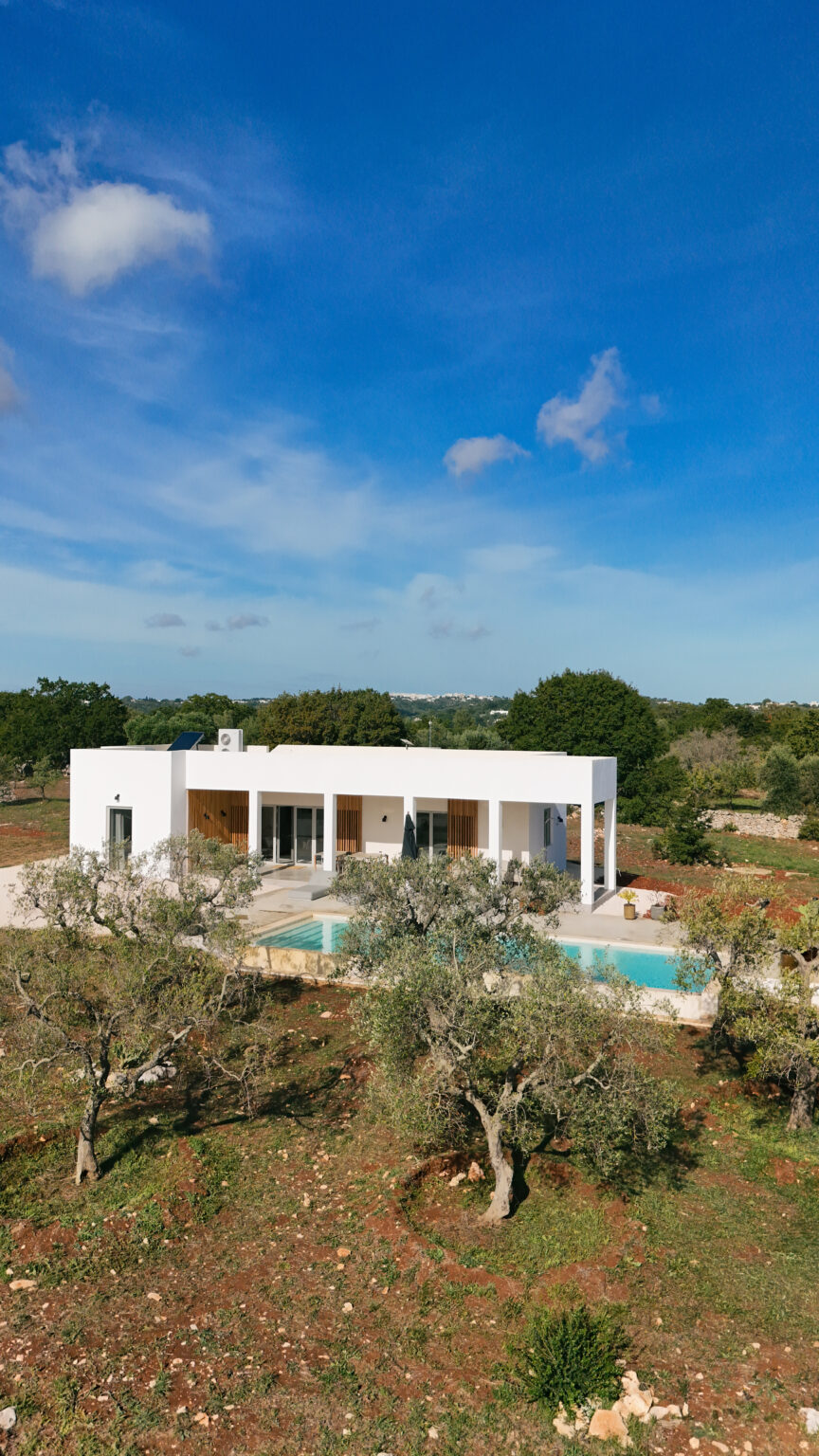 Modern white villa with a swimming pool, surrounded by lush greenery and trees, set against a clear blue sky. Ideal for luxury living or vacation rental. | Only on SopranoVillas