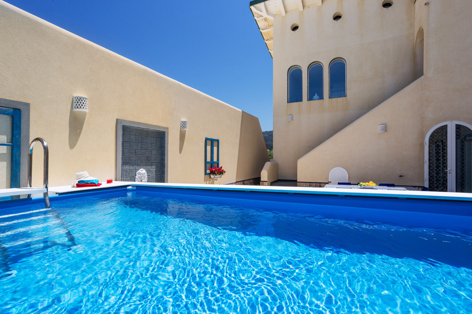 Modern outdoor swimming pool with crystal clear water, surrounded by a beige building featuring large windows and a sunny blue sky. Ideal for relaxation and leisure. | Only on SopranoVillas