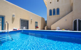 Modern outdoor swimming pool with crystal clear water, surrounded by a beige building featuring large windows and a sunny blue sky. Ideal for relaxation and leisure.