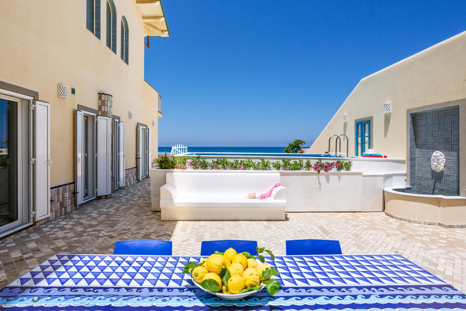 Bright outdoor terrace with a stunning ocean view, featuring a blue and white patterned dining table adorned with a bowl of lemons, a white sofa, and colorful flowers along the edge. The space is bathed in sunlight, showcasing a serene atmosphere perfect for relaxation. | Only on SopranoVillas