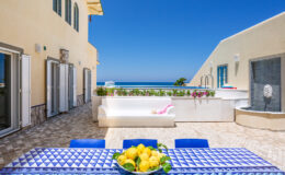 Bright outdoor terrace with a stunning ocean view, featuring a blue and white patterned dining table adorned with a bowl of lemons, a white sofa, and colorful flowers along the edge. The space is bathed in sunlight, showcasing a serene atmosphere perfect for relaxation.