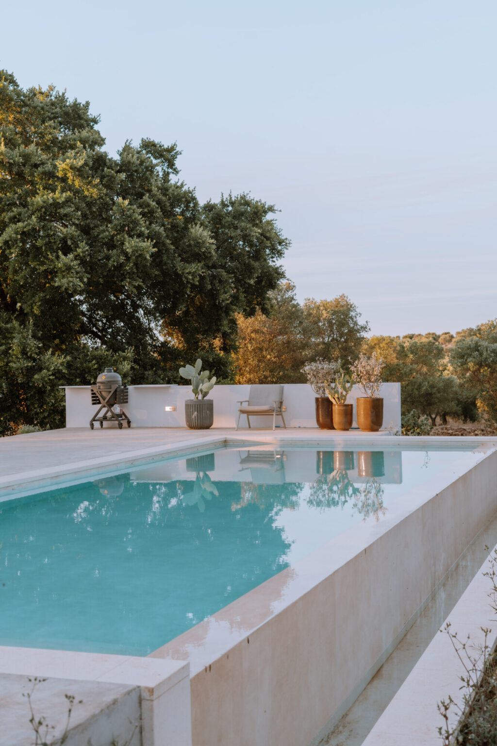 Modern outdoor pool area featuring a sleek design, surrounded by lush greenery and stylish planters. A comfortable chair and a grill are positioned nearby, creating an inviting atmosphere for relaxation and entertainment. | Only on SopranoVillas