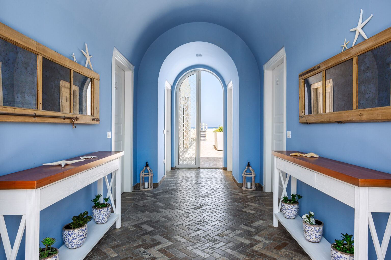 Bright and airy hallway with blue walls, featuring wooden console tables, decorative mirrors, and potted plants. The arched entrance leads to an outdoor space with a view, enhancing the coastal aesthetic. Natural light floods the space, creating a welcoming atmosphere. | Only on SopranoVillas