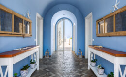 Bright and airy hallway with blue walls, featuring wooden console tables, decorative mirrors, and potted plants. The arched entrance leads to an outdoor space with a view, enhancing the coastal aesthetic. Natural light floods the space, creating a welcoming atmosphere.