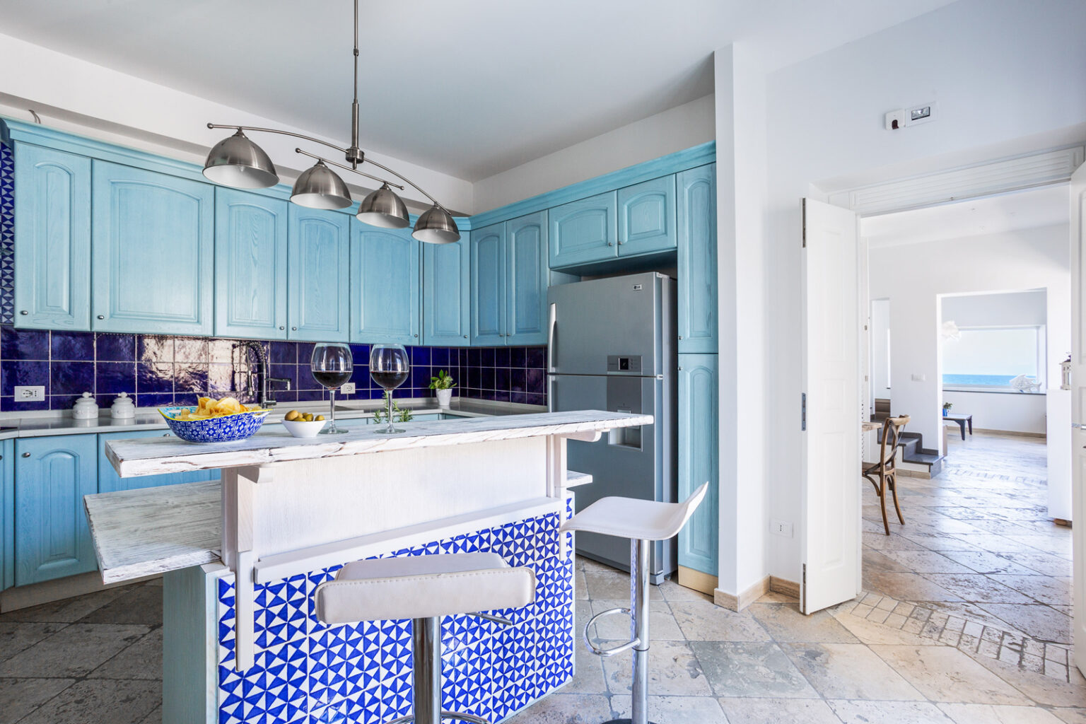 Bright and airy modern kitchen featuring blue cabinetry, a blue and white tiled island, stainless steel appliances, and an open layout leading to a scenic view. | Only on SopranoVillas