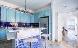 Bright and airy modern kitchen featuring blue cabinetry, a blue and white tiled island, stainless steel appliances, and an open layout leading to a scenic view.