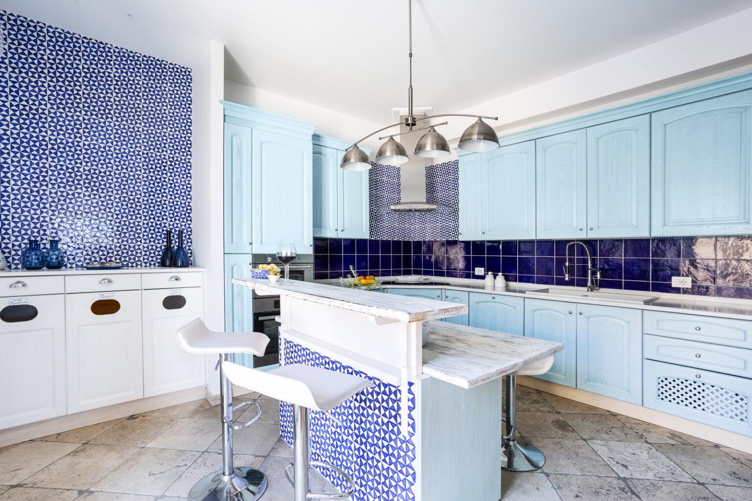 Bright and modern kitchen featuring light blue cabinetry, geometric blue tile backsplash, and a marble countertop island with bar stools. The design incorporates stylish pendant lighting and a spacious layout, ideal for cooking and entertaining. | Only on SopranoVillas