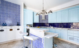 Bright and modern kitchen featuring light blue cabinetry, geometric blue tile backsplash, and a marble countertop island with bar stools. The design incorporates stylish pendant lighting and a spacious layout, ideal for cooking and entertaining.