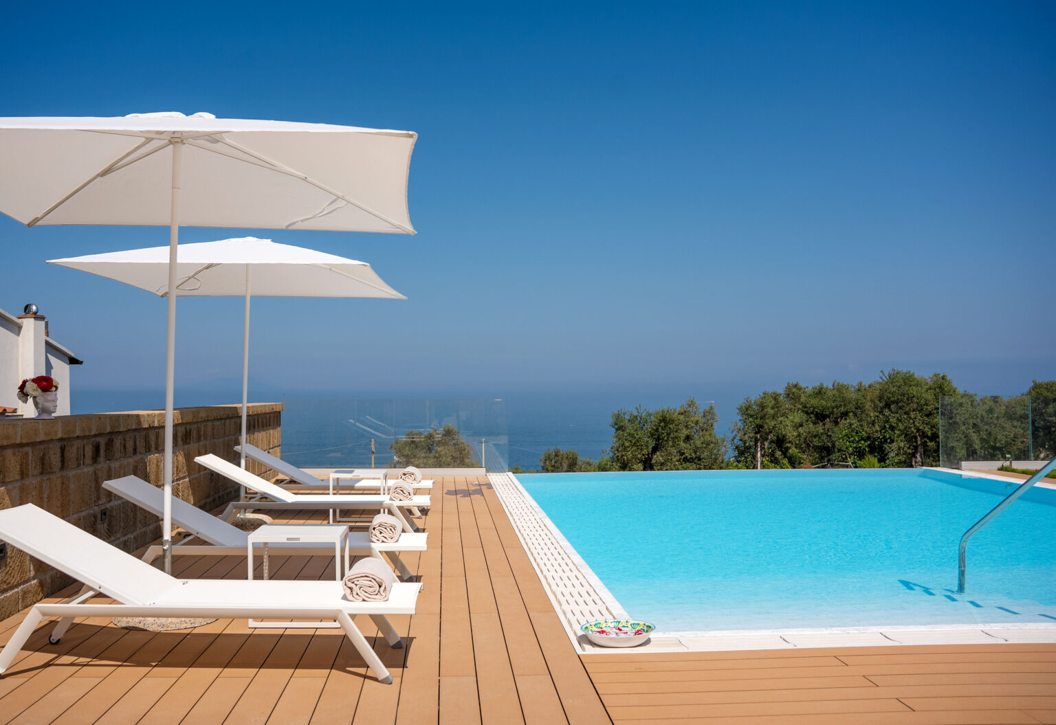 Luxurious outdoor swimming pool with sun loungers and white umbrellas, overlooking a serene ocean view and surrounded by lush greenery, ideal for relaxation and leisure. | Only on SopranoVillas