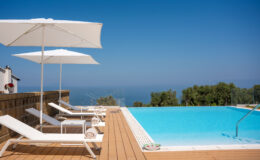 Luxurious outdoor swimming pool with sun loungers and white umbrellas, overlooking a serene ocean view and surrounded by lush greenery, ideal for relaxation and leisure.