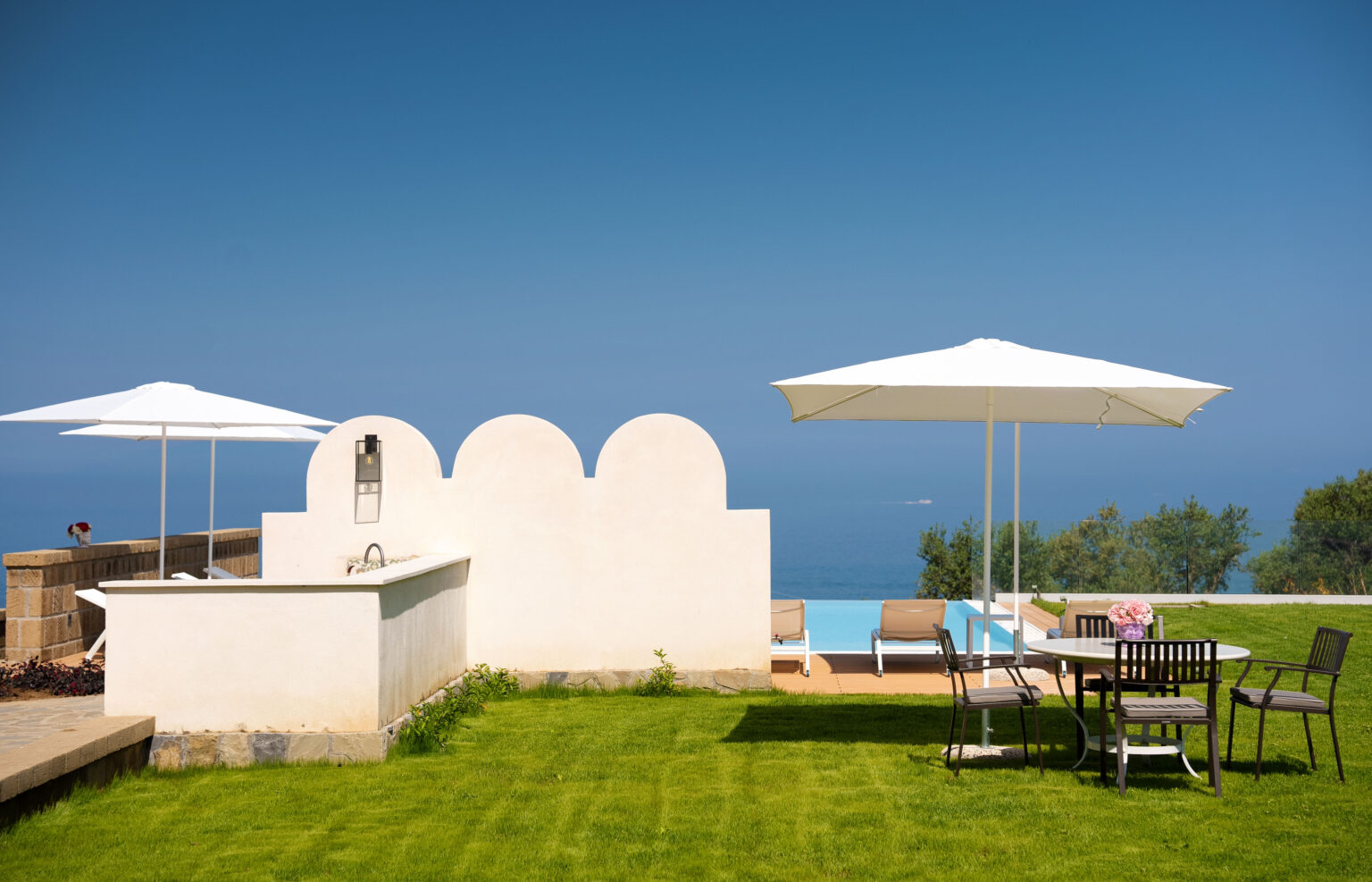 Luxurious outdoor patio featuring a well-manicured lawn, sun loungers under white umbrellas, and a dining area with a scenic view of the ocean. Ideal for relaxation and outdoor entertaining. | Only on SopranoVillas