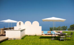 Luxurious outdoor patio featuring a well-manicured lawn, sun loungers under white umbrellas, and a dining area with a scenic view of the ocean. Ideal for relaxation and outdoor entertaining.