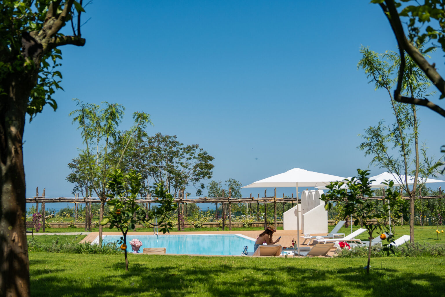 Tranquil outdoor pool area surrounded by lush greenery, featuring sun loungers and umbrellas, with a clear blue sky and distant ocean view. Ideal for relaxation and leisure in a scenic setting. | Only on SopranoVillas