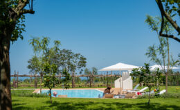 Tranquil outdoor pool area surrounded by lush greenery, featuring sun loungers and umbrellas, with a clear blue sky and distant ocean view. Ideal for relaxation and leisure in a scenic setting.