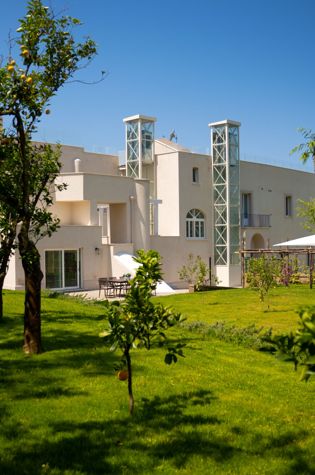 Modern residential building with a glass elevator, surrounded by lush greenery and a well-maintained garden, featuring outdoor seating and a sunny atmosphere. | Only on SopranoVillas