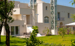 Modern residential building with a glass elevator, surrounded by lush greenery and a well-maintained garden, featuring outdoor seating and a sunny atmosphere.