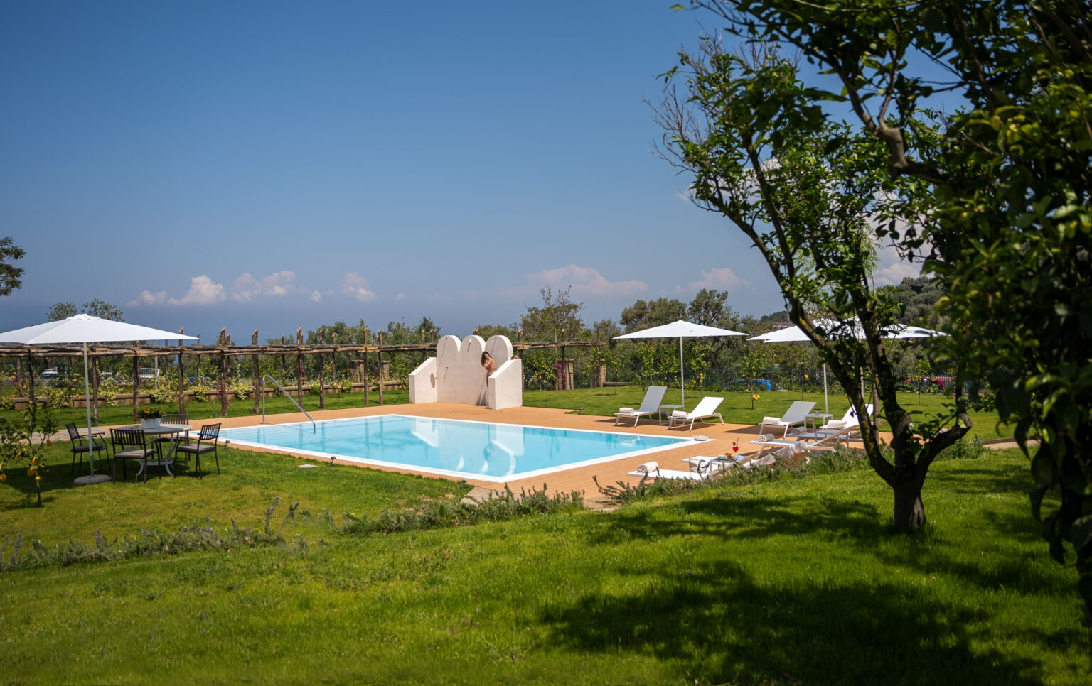 Luxury outdoor swimming pool surrounded by lush greenery and sun loungers, with umbrellas providing shade, set against a clear blue sky. Ideal for relaxation and leisure. | Only on SopranoVillas