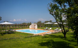 Luxury outdoor swimming pool surrounded by lush greenery and sun loungers, with umbrellas providing shade, set against a clear blue sky. Ideal for relaxation and leisure.