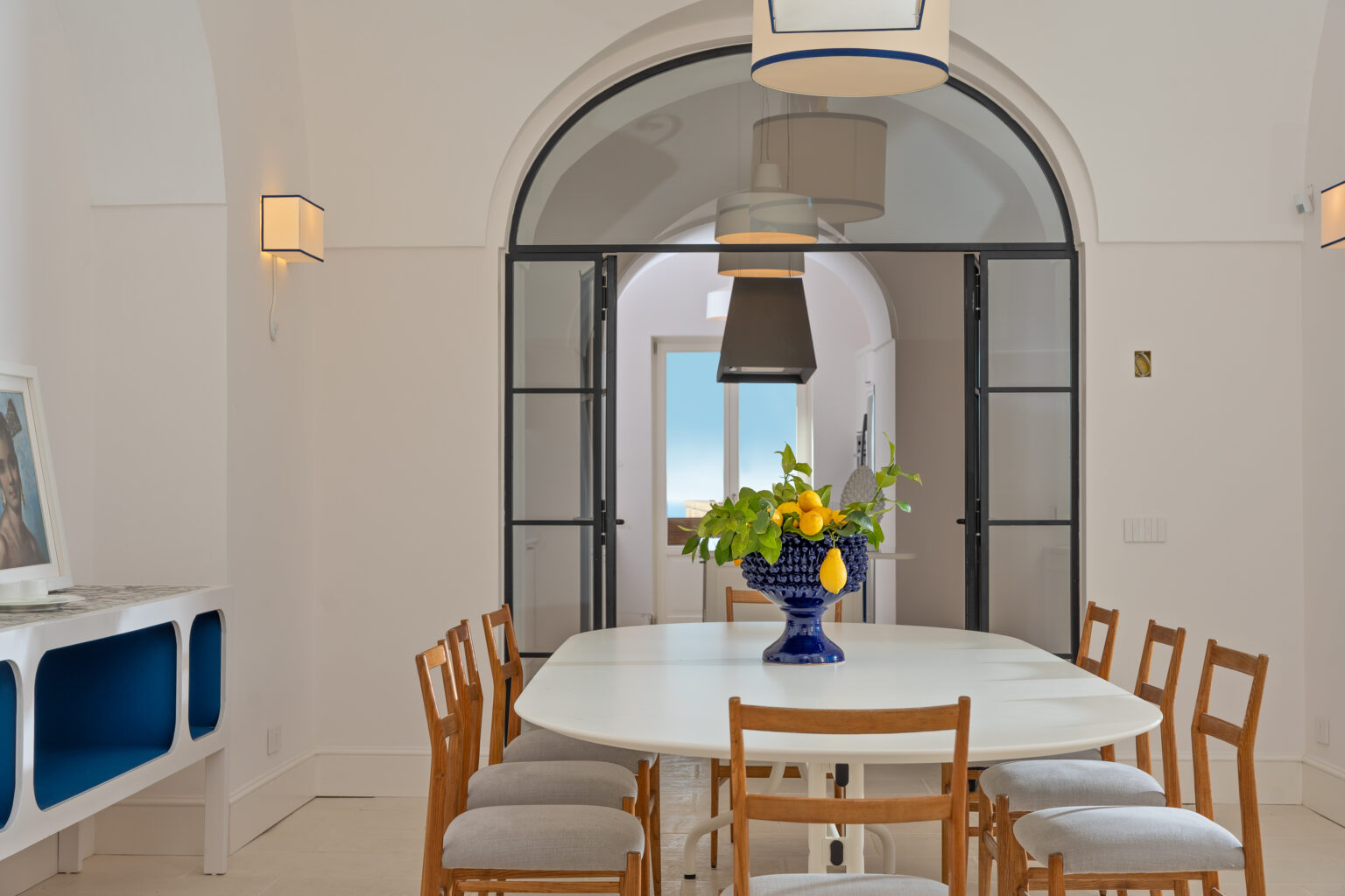 Bright and airy dining room featuring a large white table surrounded by wooden chairs, a colorful floral centerpiece, and modern pendant lighting, with an arched doorway leading to a scenic view. | Only on SopranoVillas