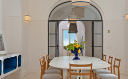 Bright and airy dining room featuring a large white table surrounded by wooden chairs, a colorful floral centerpiece, and modern pendant lighting, with an arched doorway leading to a scenic view.