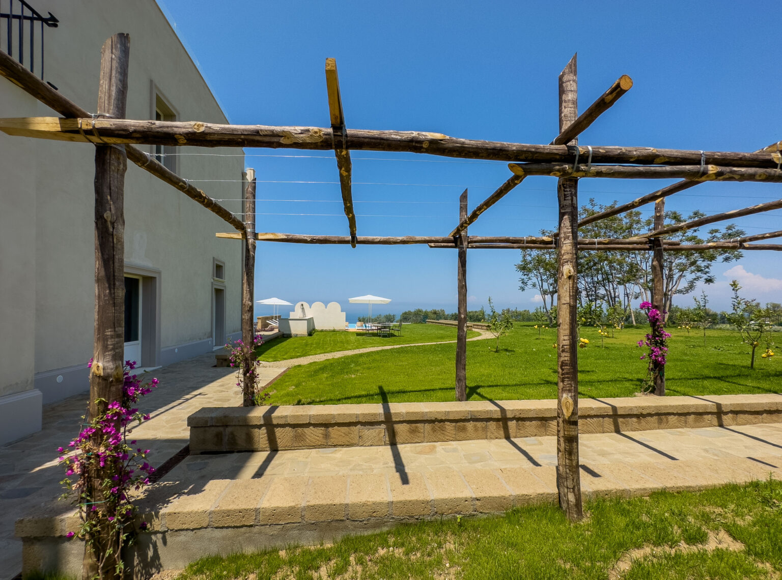 Scenic view of a landscaped garden featuring a wooden pergola, vibrant flowers, and sun loungers under umbrellas, with a clear blue sky in the background. | Only on SopranoVillas