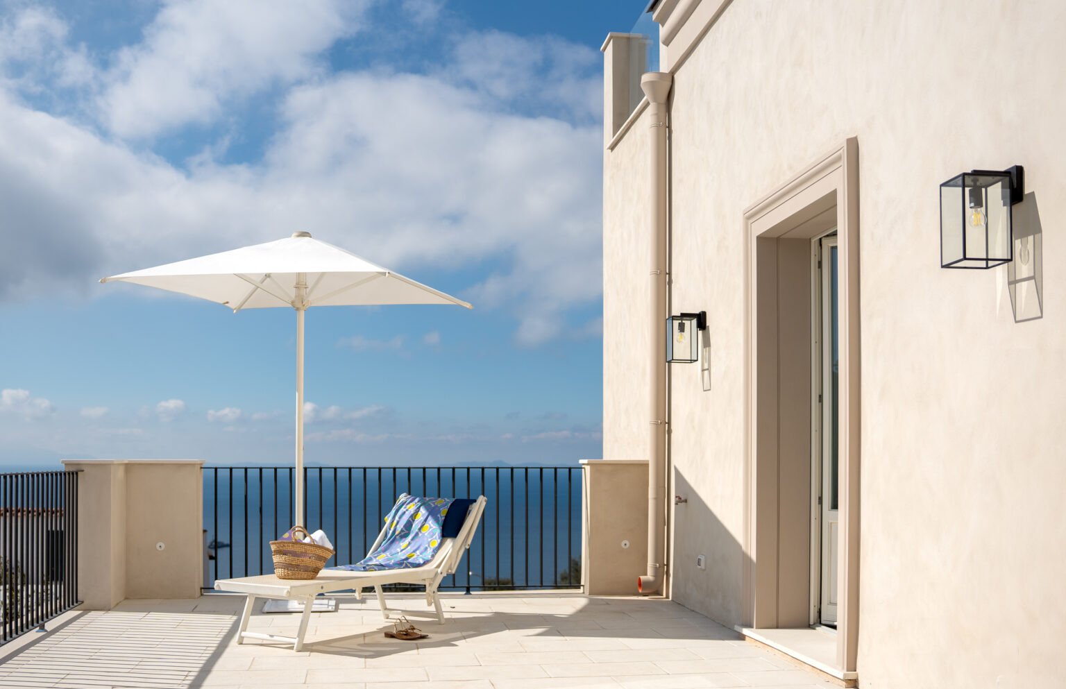 Cozy outdoor terrace featuring a sun lounger and a large umbrella, overlooking a scenic view of the ocean under a blue sky with clouds. Ideal for relaxation and enjoying coastal breezes. | Only on SopranoVillas