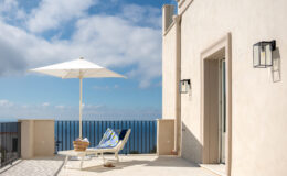Cozy outdoor terrace featuring a sun lounger and a large umbrella, overlooking a scenic view of the ocean under a blue sky with clouds. Ideal for relaxation and enjoying coastal breezes.