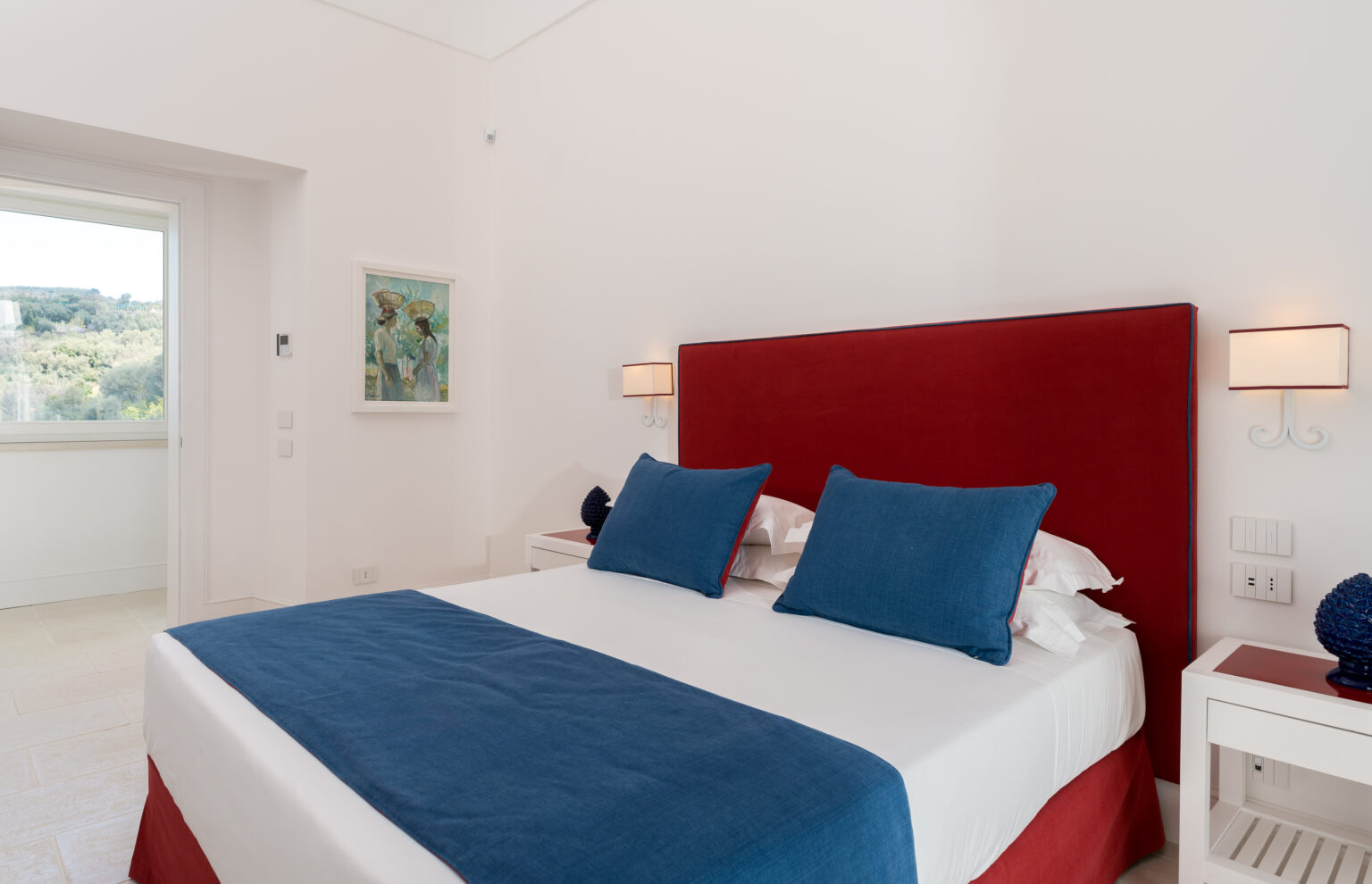 Modern bedroom featuring a red upholstered headboard, blue accent pillows, and white bedding, with natural light streaming in from a nearby window. | Only on SopranoVillas