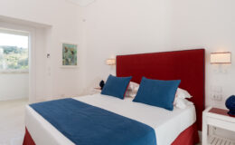 Modern bedroom featuring a red upholstered headboard, blue accent pillows, and white bedding, with natural light streaming in from a nearby window.
