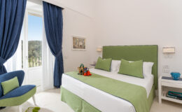 Bright and modern hotel bedroom featuring a green bedspread, blue curtains, and stylish decor, with a view of nature through the window. Ideal for a relaxing getaway.