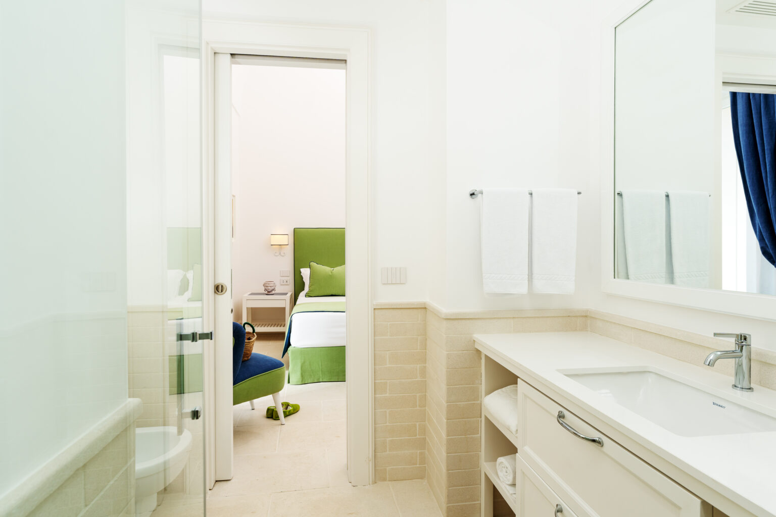 Modern bathroom with a sleek vanity and large mirror, featuring a glass door leading to a cozy bedroom with green accents and a bright atmosphere. | Only on SopranoVillas
