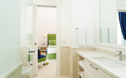 Modern bathroom with a sleek vanity and large mirror, featuring a glass door leading to a cozy bedroom with green accents and a bright atmosphere.