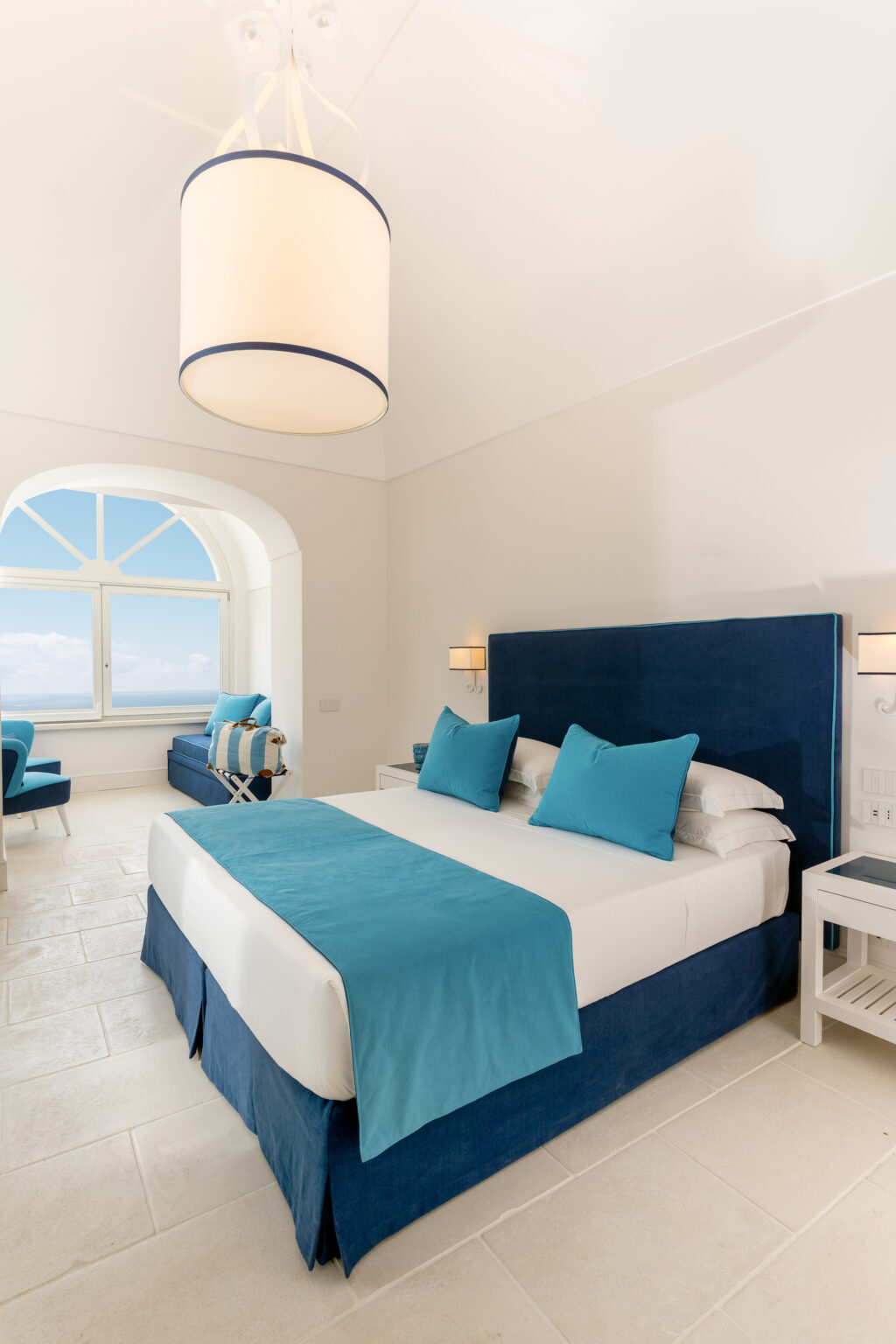 Luxurious hotel room featuring a blue and white color scheme, with a comfortable bed adorned with turquoise accents, a stylish pendant light, and a scenic window view overlooking the ocean. | Only on SopranoVillas