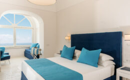 Luxurious hotel room featuring a blue and white color scheme, with a comfortable bed adorned with turquoise accents, a stylish pendant light, and a scenic window view overlooking the ocean.