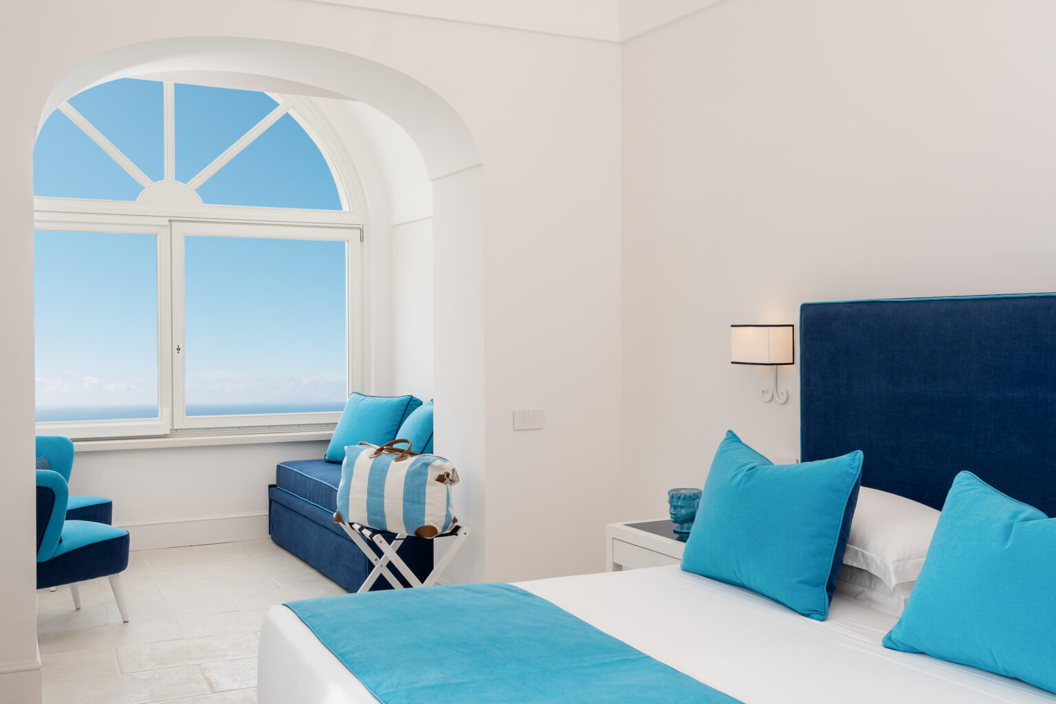 Bright and modern hotel room featuring a blue and white color scheme, with a large window showcasing a clear sky and ocean view. The room includes a comfortable bed with decorative pillows, a cozy seating area, and stylish furnishings, perfect for a relaxing getaway. | Only on SopranoVillas
