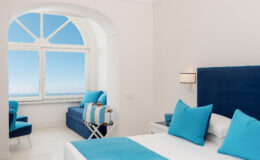 Bright and modern hotel room featuring a blue and white color scheme, with a large window showcasing a clear sky and ocean view. The room includes a comfortable bed with decorative pillows, a cozy seating area, and stylish furnishings, perfect for a relaxing getaway.