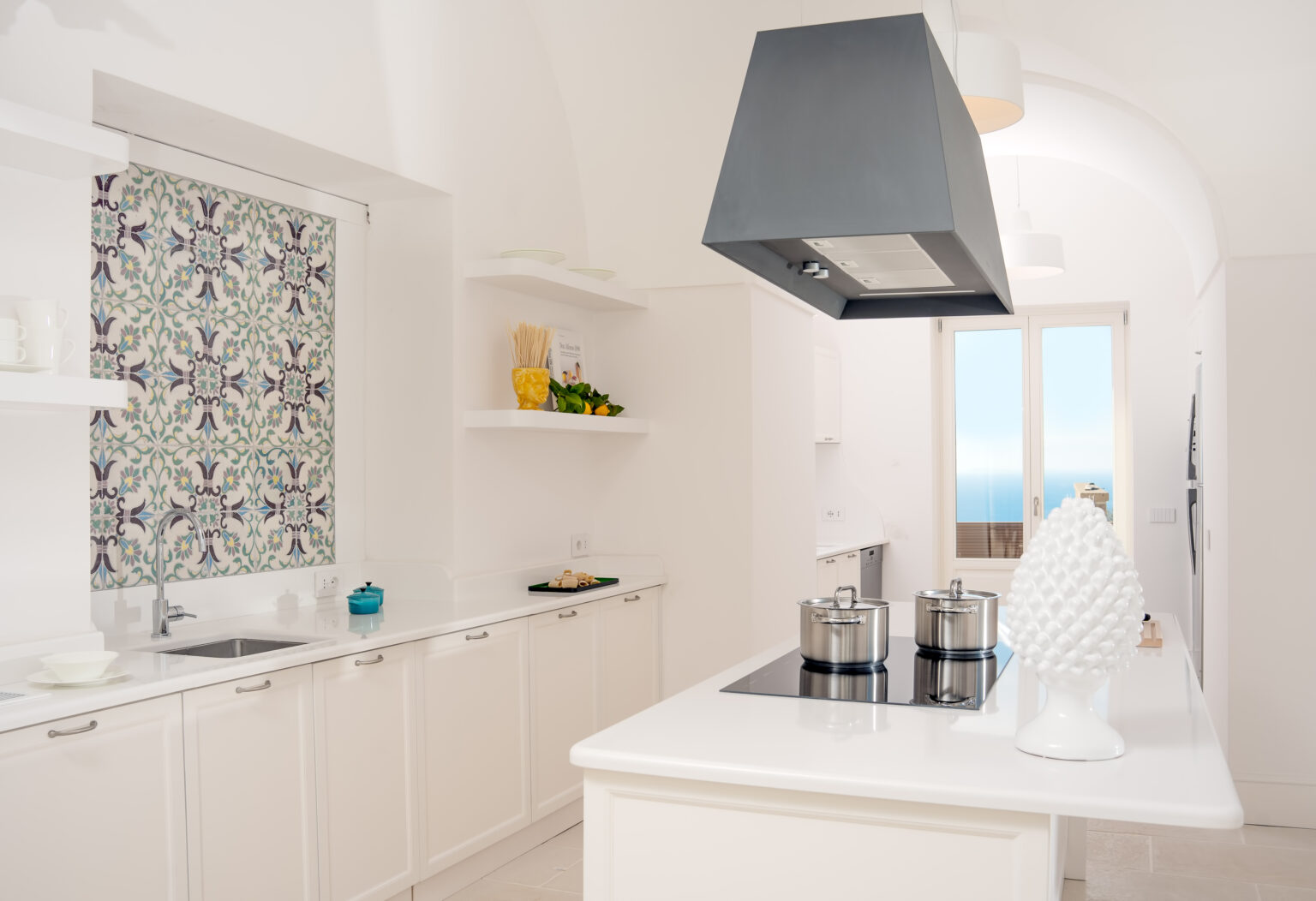 Modern kitchen featuring white cabinetry, a stylish range hood, and a vibrant patterned window treatment, with a scenic ocean view visible through large windows. The kitchen is equipped with stainless steel pots on a sleek cooktop, complemented by decorative elements like a white sculpture and fresh produce on the shelves. | Only on SopranoVillas