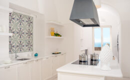 Modern kitchen featuring white cabinetry, a stylish range hood, and a vibrant patterned window treatment, with a scenic ocean view visible through large windows. The kitchen is equipped with stainless steel pots on a sleek cooktop, complemented by decorative elements like a white sculpture and fresh produce on the shelves.
