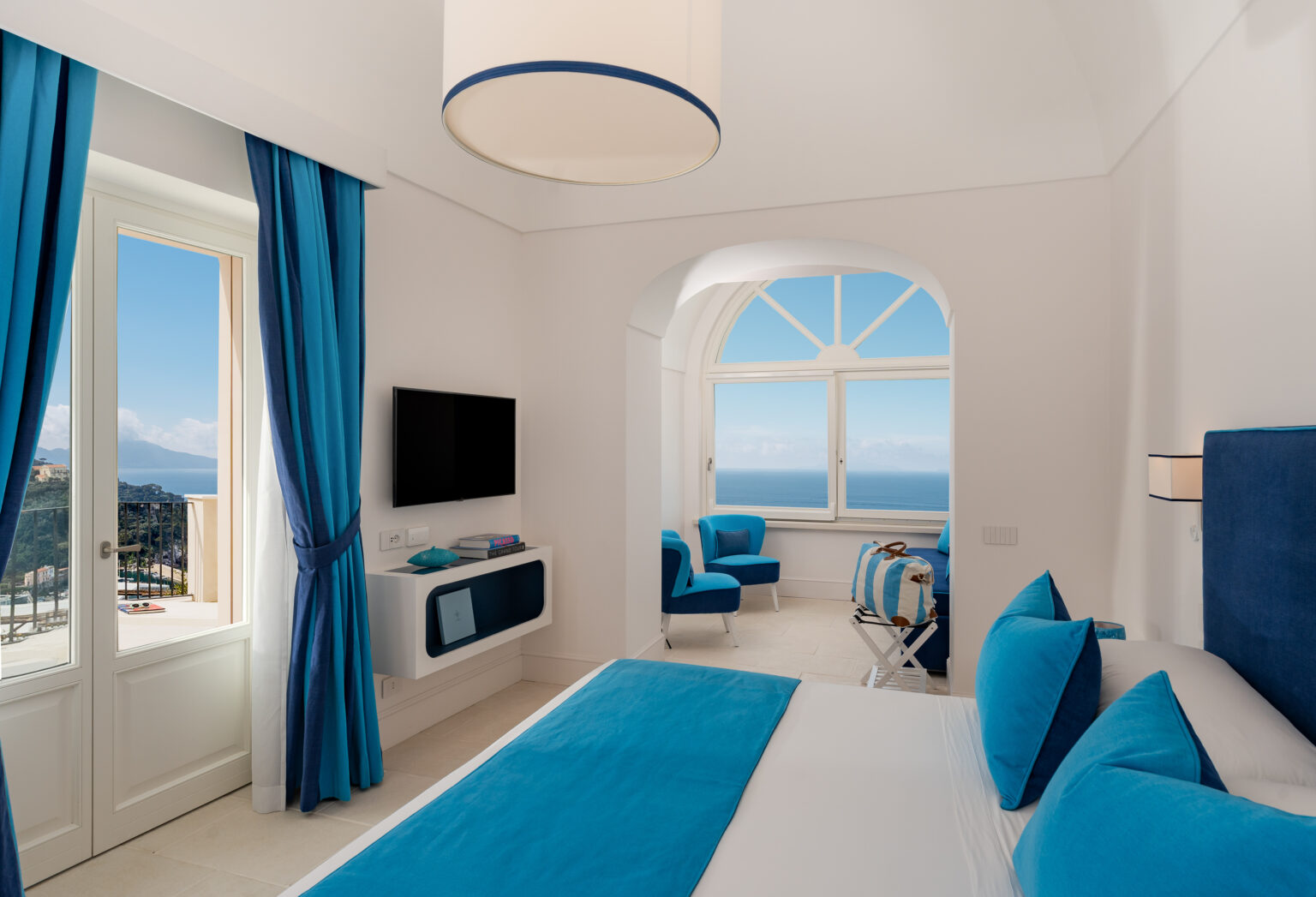 Modern hotel room with blue and white decor, featuring a large window with ocean views, a flat-screen TV, and stylish seating. Ideal for a relaxing getaway. | Only on SopranoVillas