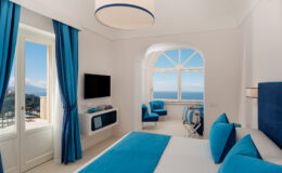 Modern hotel room with blue and white decor, featuring a large window with ocean views, a flat-screen TV, and stylish seating. Ideal for a relaxing getaway.