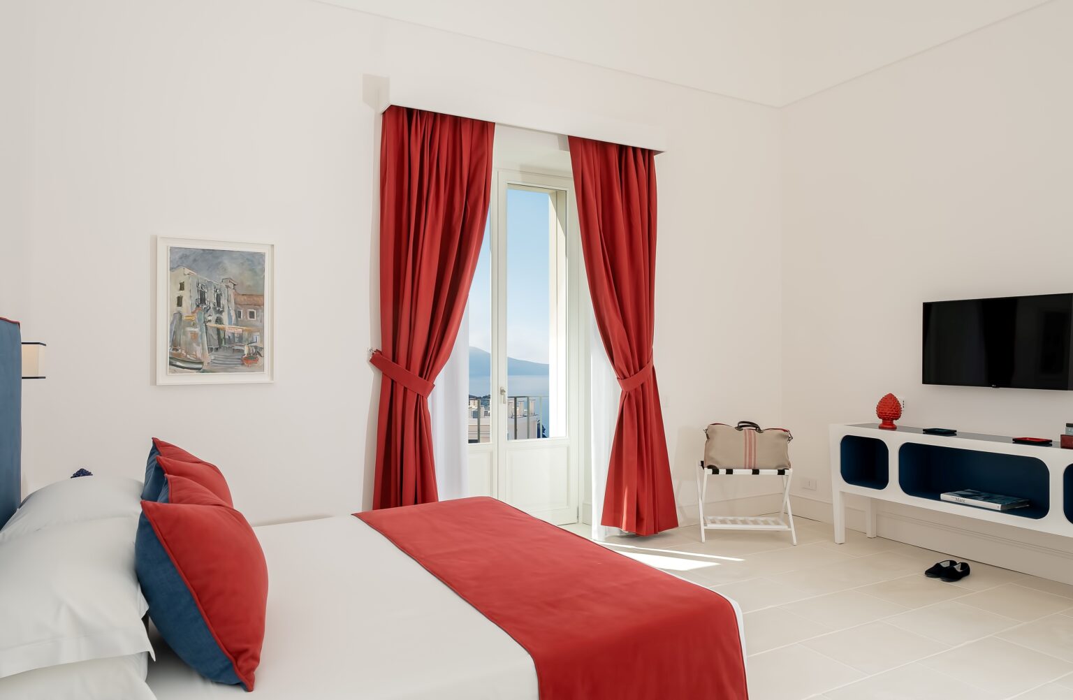 Modern bedroom with white walls, a red and blue color scheme, large windows with red curtains, a flat-screen TV, and a stylish side table. Ideal for relaxation and comfort. | Only on SopranoVillas