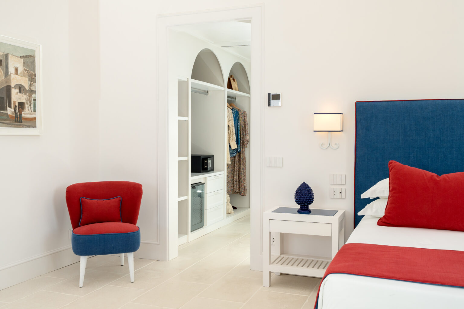 Modern bedroom interior featuring a blue and red color scheme, with a comfortable bed, stylish side table, and a cozy armchair. A closet is visible in the background, showcasing organized storage with a microwave. Natural light enhances the bright, airy atmosphere of the space. | Only on SopranoVillas