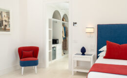 Modern bedroom interior featuring a blue and red color scheme, with a comfortable bed, stylish side table, and a cozy armchair. A closet is visible in the background, showcasing organized storage with a microwave. Natural light enhances the bright, airy atmosphere of the space.