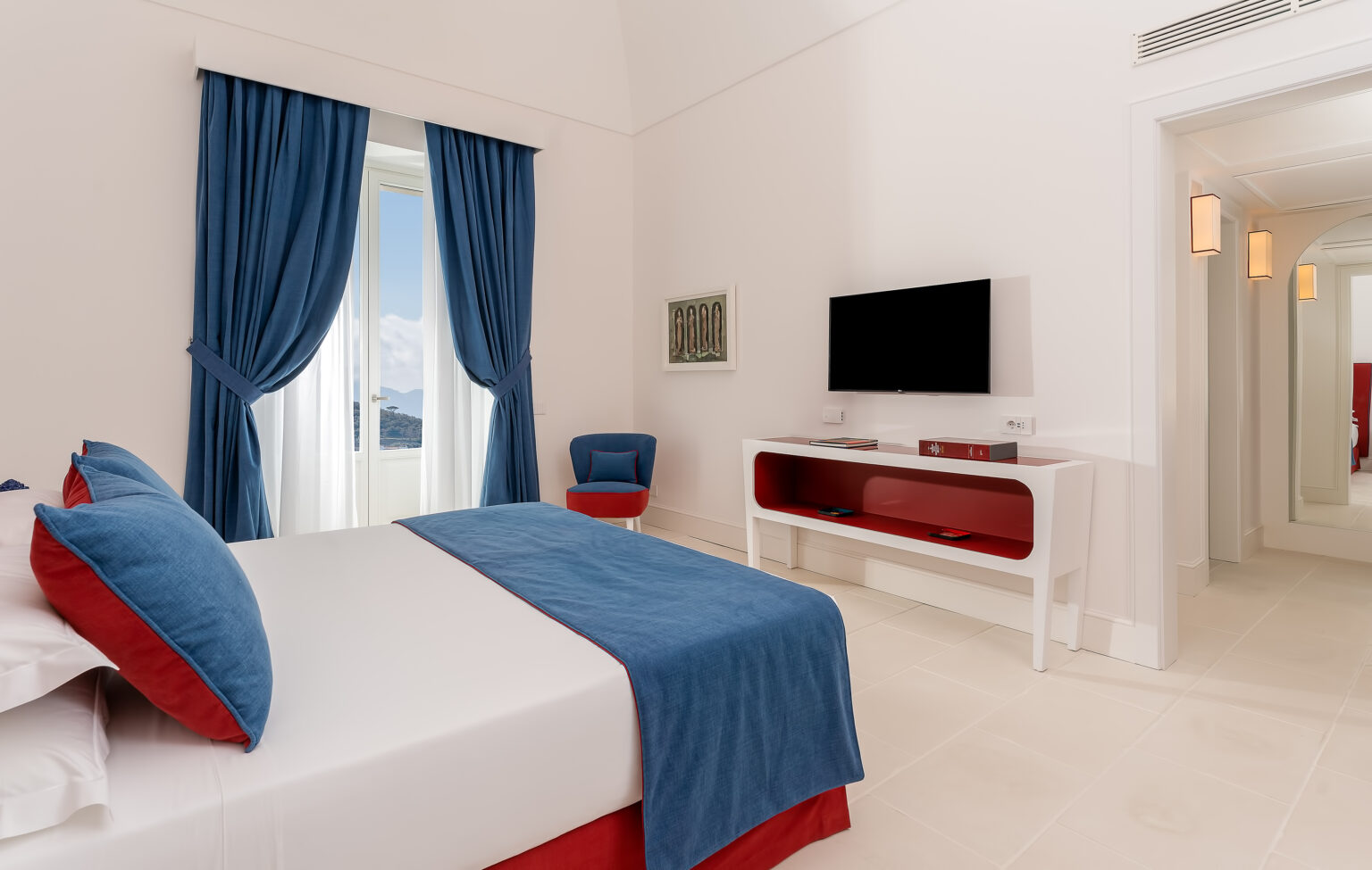 Modern hotel room featuring a double bed with blue and red accents, a flat-screen TV, and large windows with blue curtains, creating a bright and inviting atmosphere. | Only on SopranoVillas
