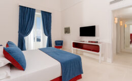 Modern hotel room featuring a double bed with blue and red accents, a flat-screen TV, and large windows with blue curtains, creating a bright and inviting atmosphere.