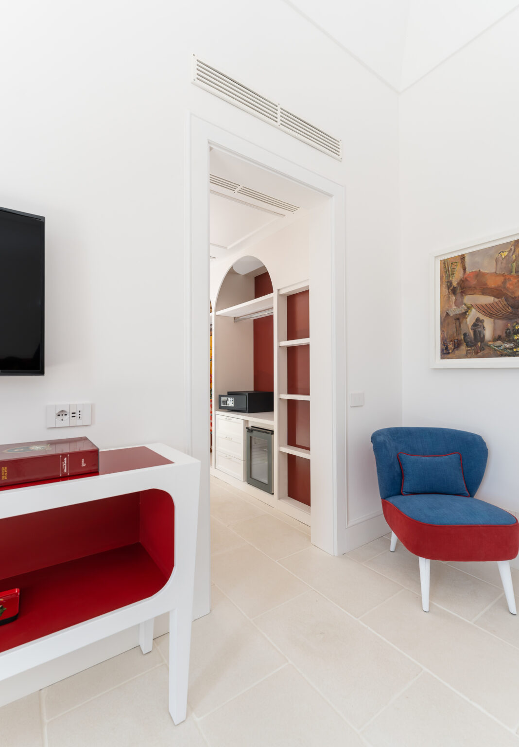 Modern interior design featuring a stylish living area with a white console table, a blue and red accent chair, and a doorway leading to a kitchenette. The space is bright and airy, showcasing clean lines, minimalist decor, and a flat-screen TV mounted on the wall. | Only on SopranoVillas