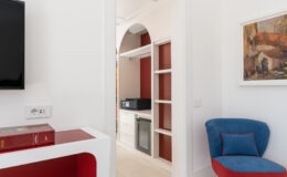 Modern interior design featuring a stylish living area with a white console table, a blue and red accent chair, and a doorway leading to a kitchenette. The space is bright and airy, showcasing clean lines, minimalist decor, and a flat-screen TV mounted on the wall.
