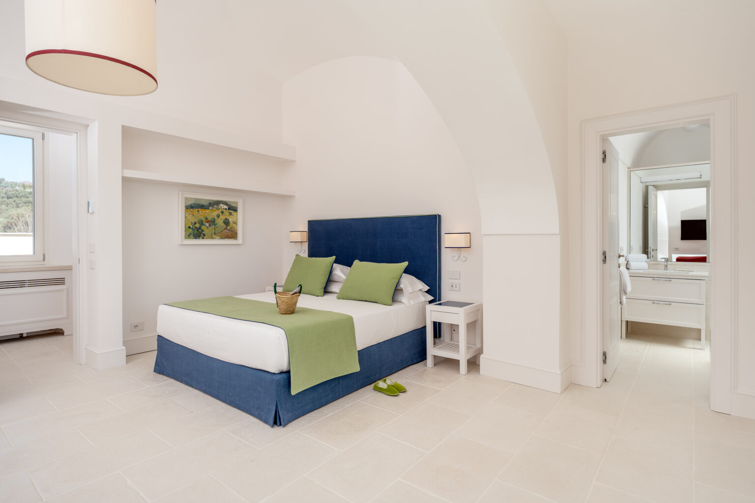 Modern bedroom featuring a blue bed with green accents, bright natural light from a window, and a stylish bathroom visible through an open door. Ideal for a relaxing stay. | Only on SopranoVillas