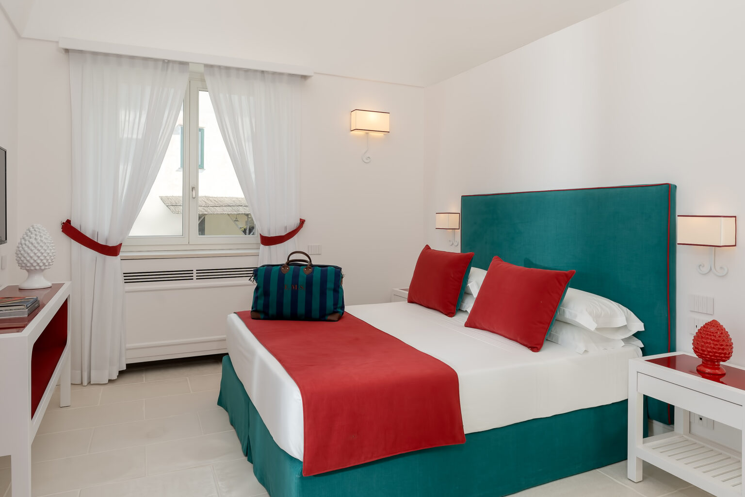 Modern hotel bedroom featuring a turquoise headboard, white bedding, and bright red accent pillows. The room includes a stylish nightstand and a decorative lamp, with sheer white curtains framing a window that allows natural light. A designer bag is placed on the bed, adding a touch of luxury to the space. | Only on SopranoVillas