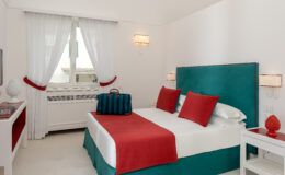 Modern hotel bedroom featuring a turquoise headboard, white bedding, and bright red accent pillows. The room includes a stylish nightstand and a decorative lamp, with sheer white curtains framing a window that allows natural light. A designer bag is placed on the bed, adding a touch of luxury to the space.